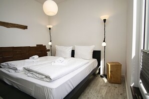 A neatly made bed with white linens and towels, a wooden headboard, two bedside lamps, and a small wooden nightstand.