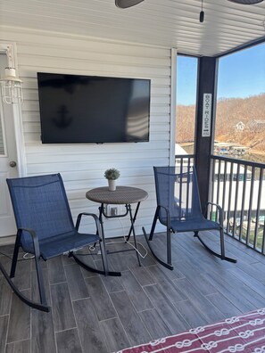 A covered outdoor seating area with two rocking chairs, a table, and a flat-screen TV mounted on the wall. - NEW LISTING *Lakefront* Sleeps 6* Pool* (Lake Ozark)