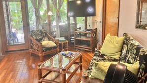 Una habitación con piso de madera, un sofá estampado con hojas, una mesa de centro de madera, un televisor y una ventana con cortinas.