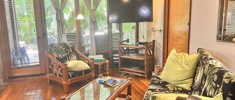 Une pièce avec un plancher en bois, un canapé orné d’un motif à feuilles, une table basse en bois, un téléviseur et une fenêtre avec des rideaux.