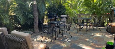 Un patio al aire libre con mobiliario de mimbre, suelo de piedra y una zona de barbacoa rodeada de abundante vegetación.