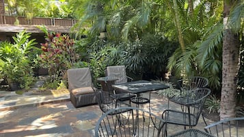 Un patio al aire libre con suelo de piedra, mobiliario de mimbre y abundante vegetación.