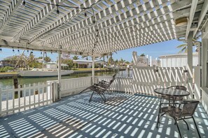 Un espacio al aire libre cubierto con una pérgola, dos sillas de metal, una pequeña mesa redonda y vistas a un puerto deportivo con barcos.