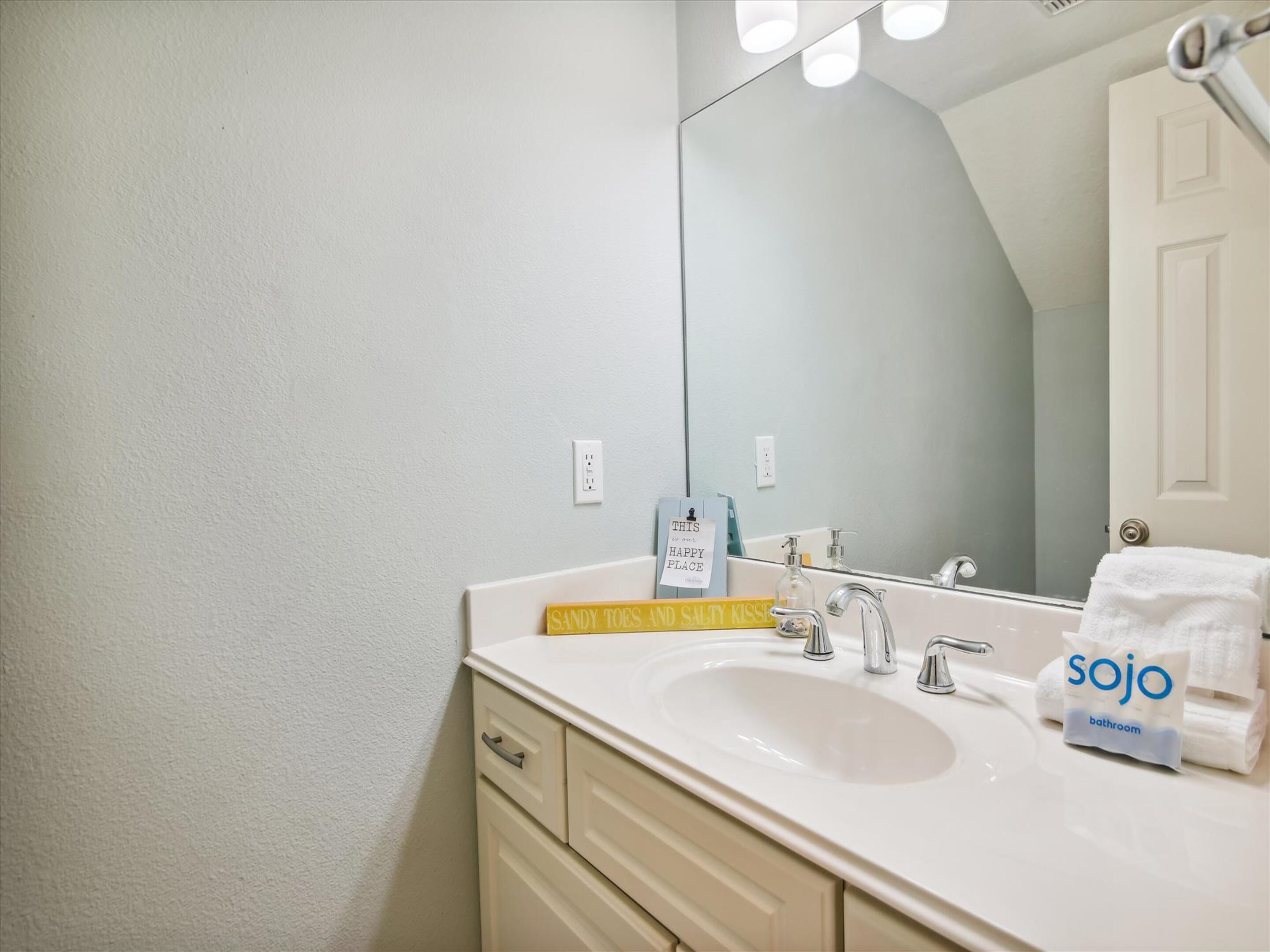 Une salle de bain avec un lavabo blanc, un miroir et un porte-serviettes. Un panneau est accroché au miroir et un sac contenant des articles de toilette est posé sur le lavabo.