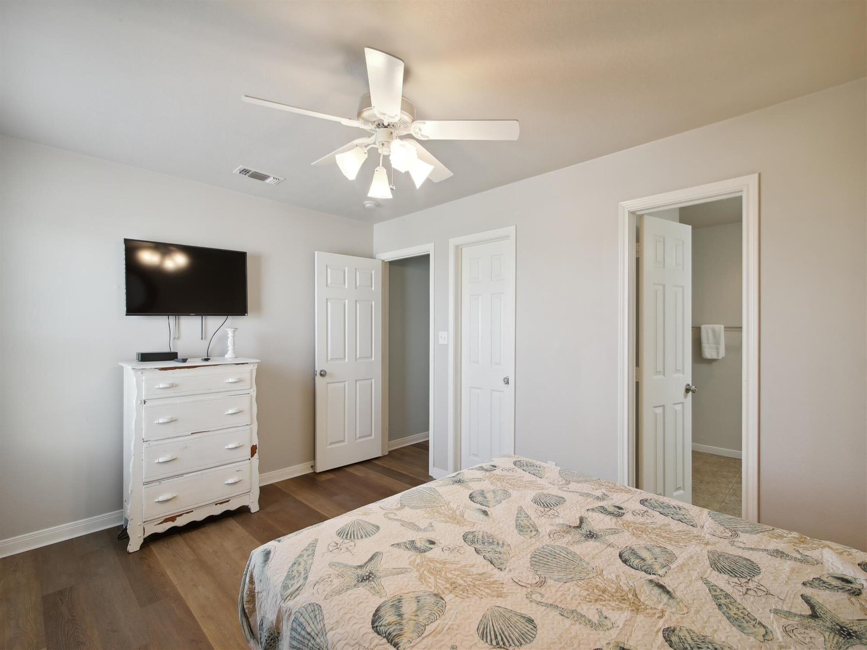 A bedroom with a bed, a dresser, a ceiling fan, a television, and two doors.