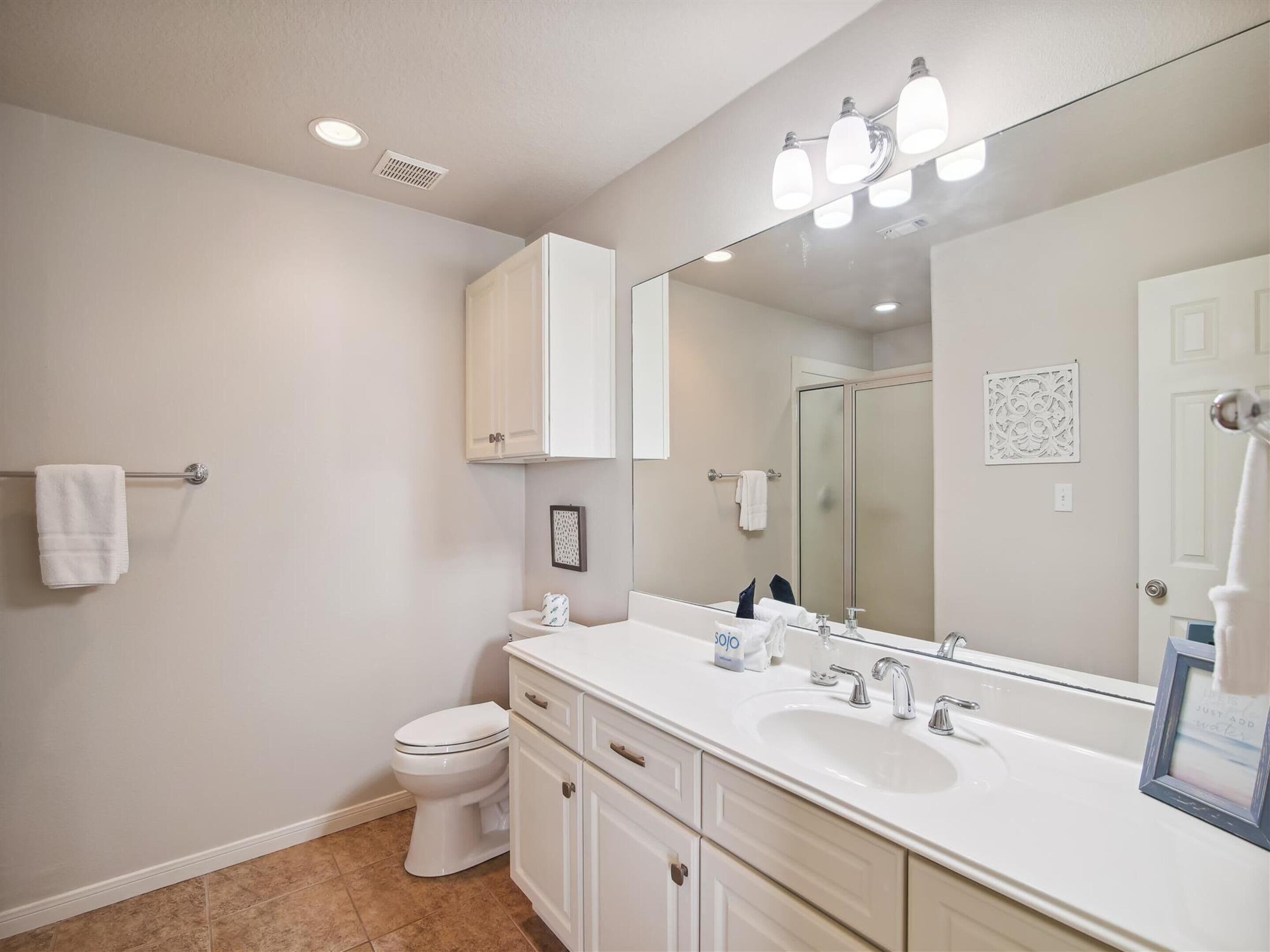 A modern bathroom with a white sink, vanity, and toilet. It features a large mirror, recessed ceiling lights, and a towel rack.