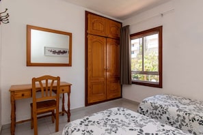 Une chambre avec un bureau en bois, une chaise, un lit avec une literie à motifs floraux, une armoire et une fenêtre donnant sur un paysage verdoyant.