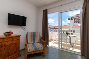 Ein Zimmer mit einem Holzsessel, einem Fernseher an der Wand, einem kleinen Tisch mit Obstschale und einem Balkon mit Tisch und Stühlen.