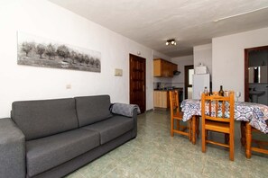 Ein Wohnzimmer mit grauem Sofa, einem Esstisch mit gemustertem Tischläufer, Holzstühlen und einer Küche im Hintergrund.