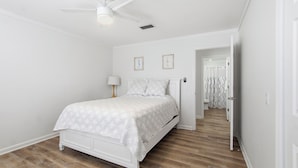 Une chambre à coucher avec un lit, un ventilateur de plafond et une salle de bain visible à travers une porte ouverte.