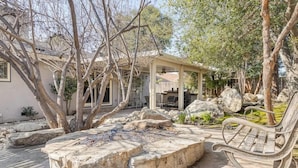 A covered outdoor seating area with a stone fire pit, surrounded by large rocks and trees.