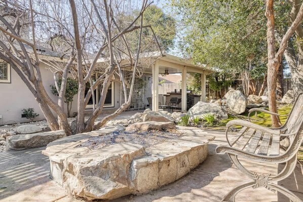 A covered outdoor seating area with a stone fire pit, surrounded by large rocks and trees.