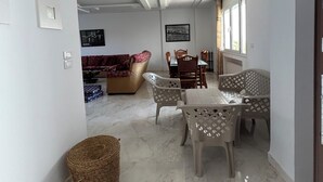 Een ruime woonkamer met een bank, eettafel en stoelen. Een televisie is aan de muur bevestigd.