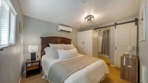 Een slaapkamer met een bed, nachtkastje, lamp, airconditioning, kledingkast en een deur naar een andere kamer.