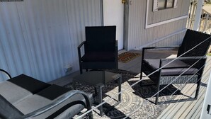 Een overdekt terras met twee loungestoelen en een kleine tafel, een kamerkleed met patroon, en een metalen hekwerk.