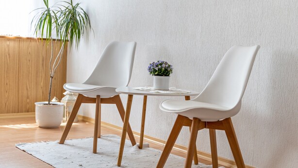 Una habitación con piso de madera, paredes blancas, un tapete blanco, dos sillas blancas, una mesita pequeña blanca y una planta en maceta.