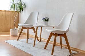 Une pièce avec un plancher en bois, des murs blancs, un tapis blanc, deux chaises blanches, une petite table blanche et une plante en pot.