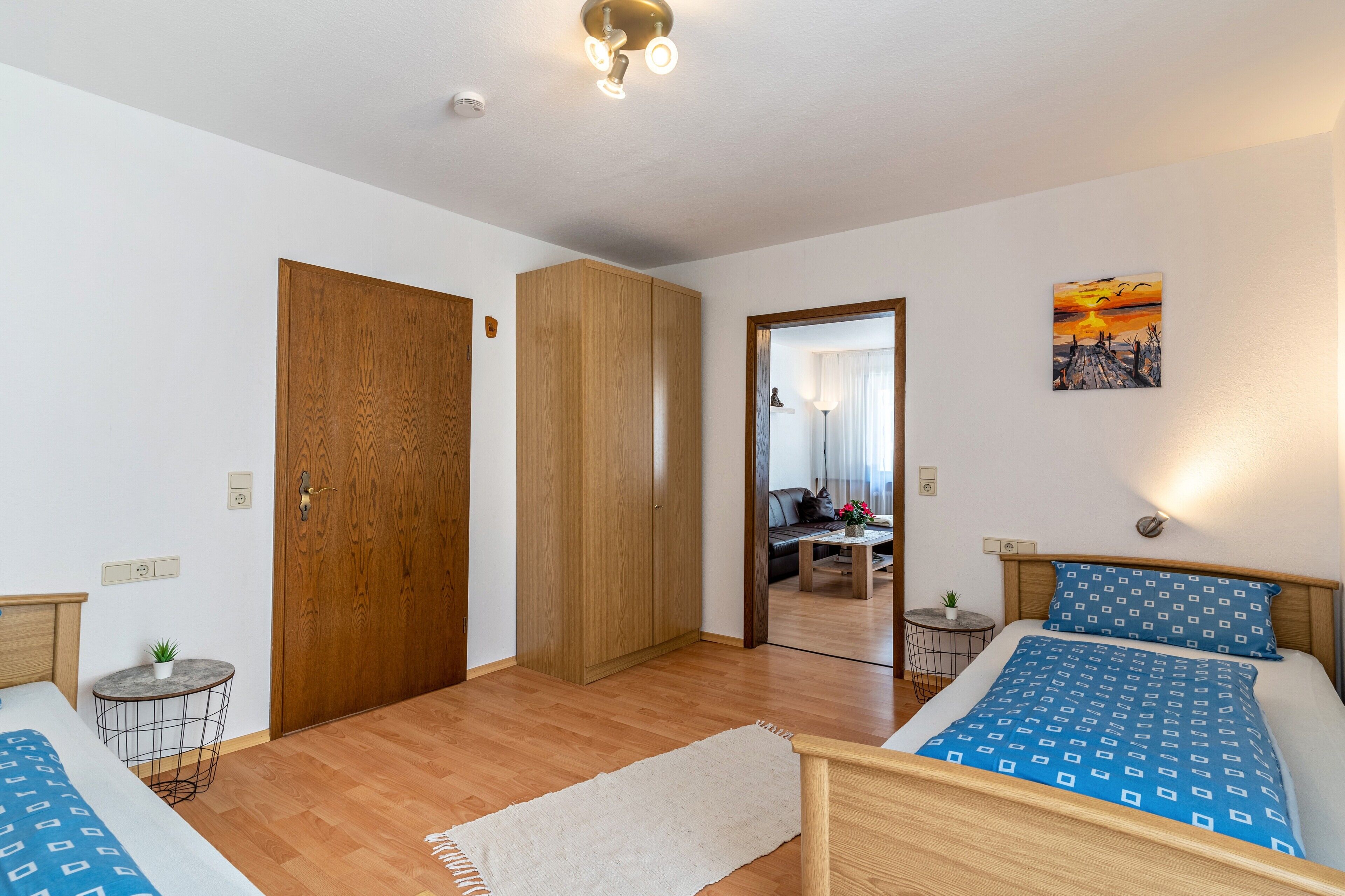 Een slaapkamer met twee bedden, een houten garderobe, een klein rond tafeltje, een spiegel en een schilderij aan de muur.