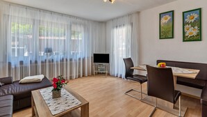Una sala de estar moderna con un sofá, una mesa de centro, una mesa de comedor y sillas. Hay un televisor de pantalla plana sobre un mueble bajo, y dos cuadros enmarcados en la pared.
