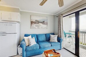 Ein Wohnzimmer mit einem blauen Sofa, einem Galanz-Kühlschrank, einem Gemälde mit Strandmotiv und einem Balkon mit Glastisch und Stühlen.