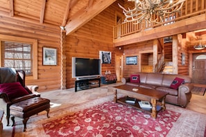 A log cabin interior with a wooden ceiling, a television, a leather sofa, and a red patterned rug.