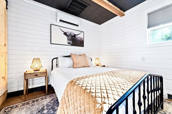A bedroom with a bed, a nightstand, a lamp, a framed picture on the wall, and a window.