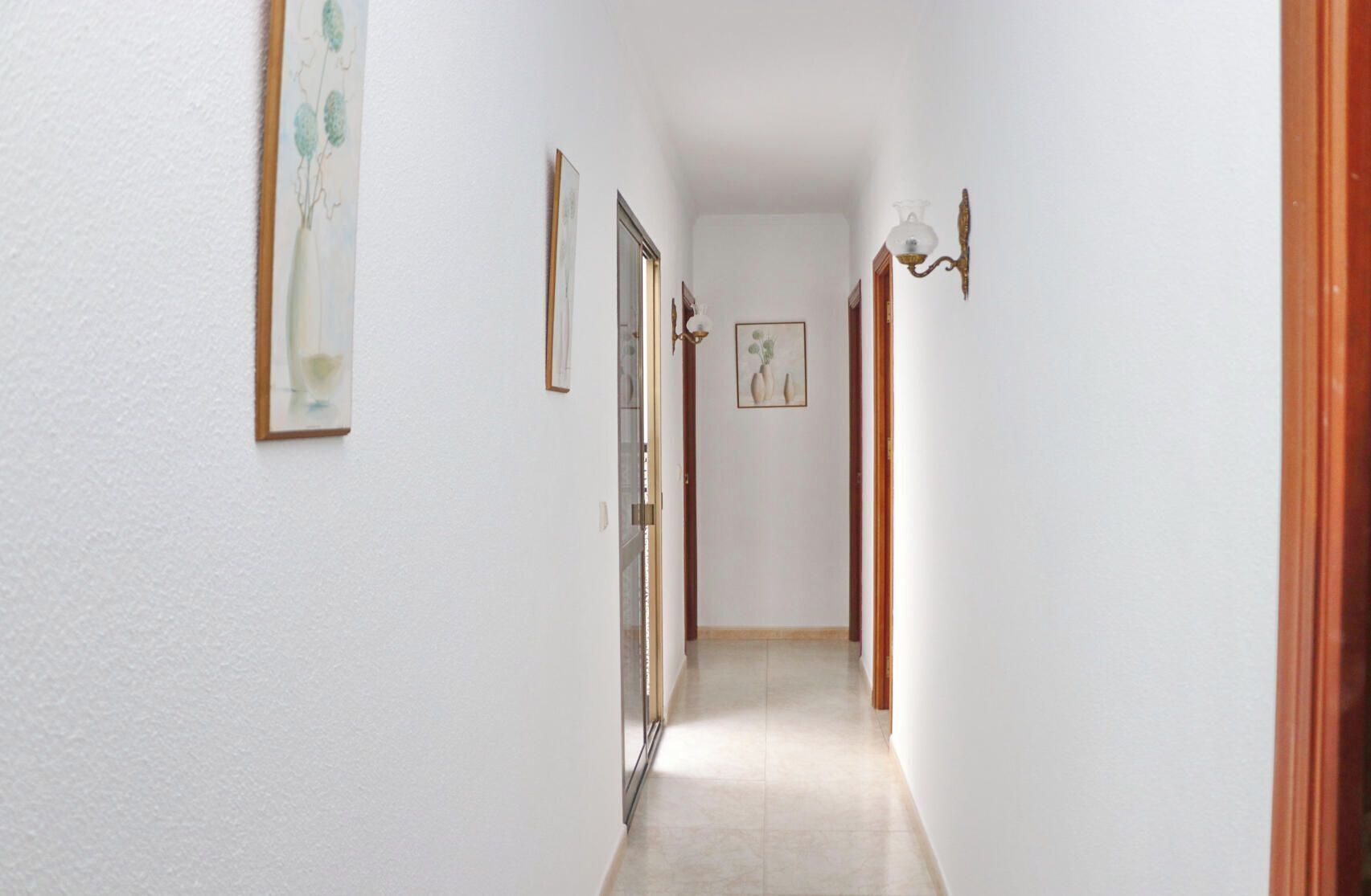 A narrow hallway with white walls, framed artwork, and a door leading to another room.