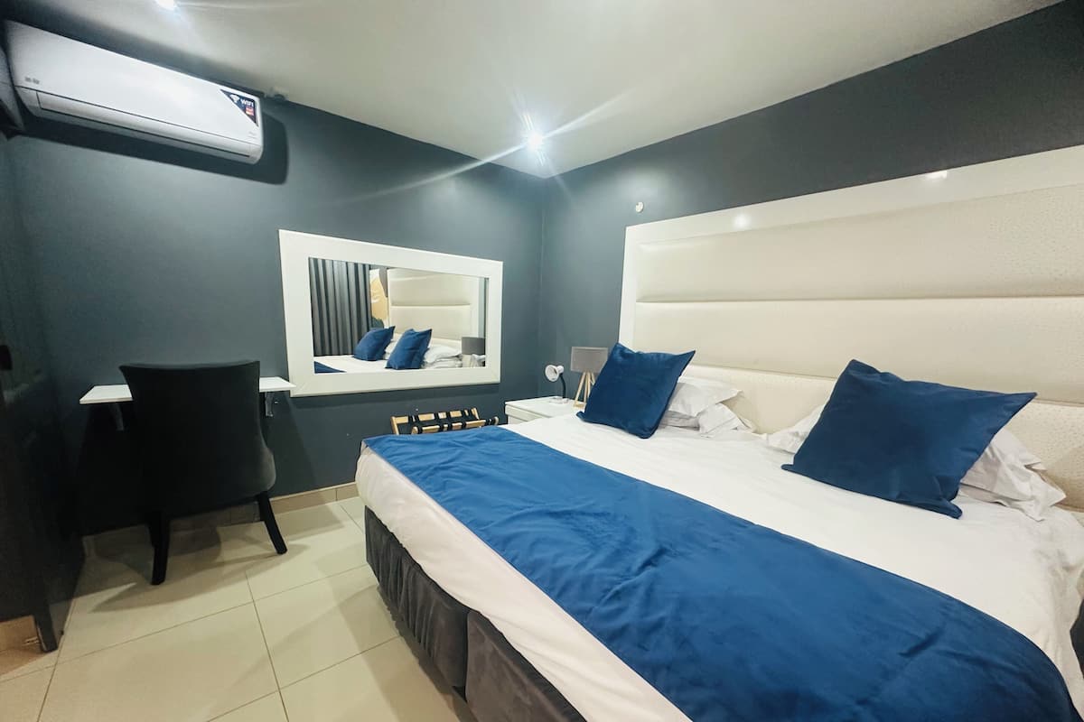 A hotel room with a bed, a desk, a chair, a mirror, and an air conditioning unit.