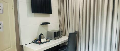 A hotel room with a flat-screen TV mounted on the wall, a microwave on the counter, a small fridge, a desk with a chair, and a curtain.