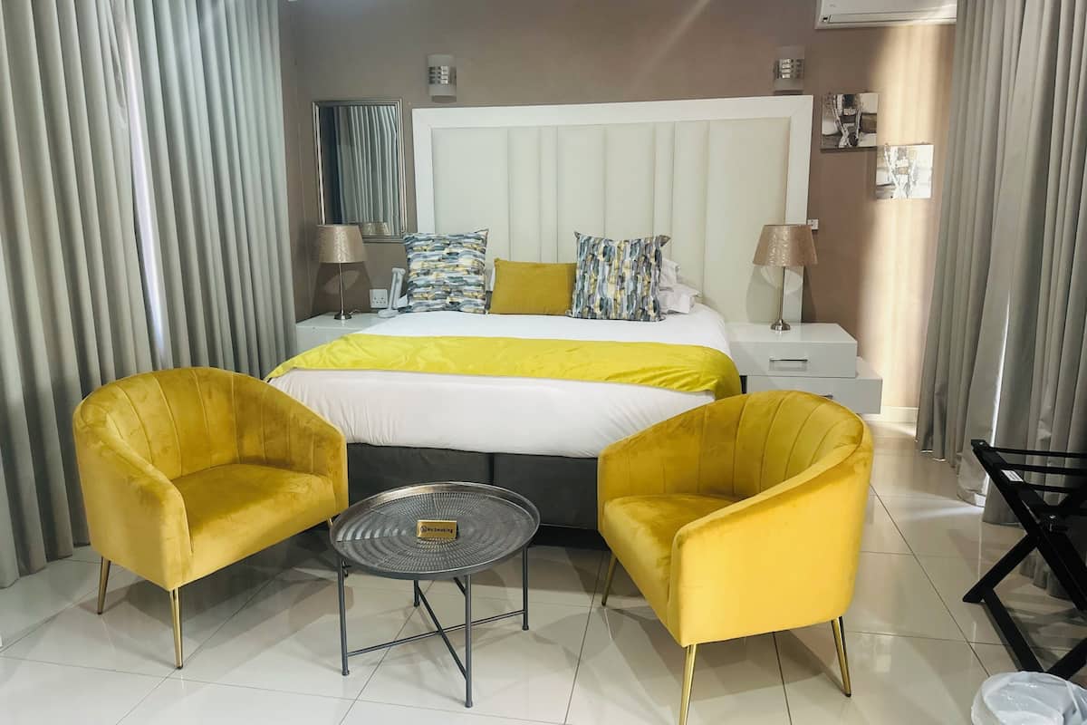 A hotel room with a large bed, two yellow armchairs, a small round table, a nightstand with a lamp, and an air conditioning unit on the wall.