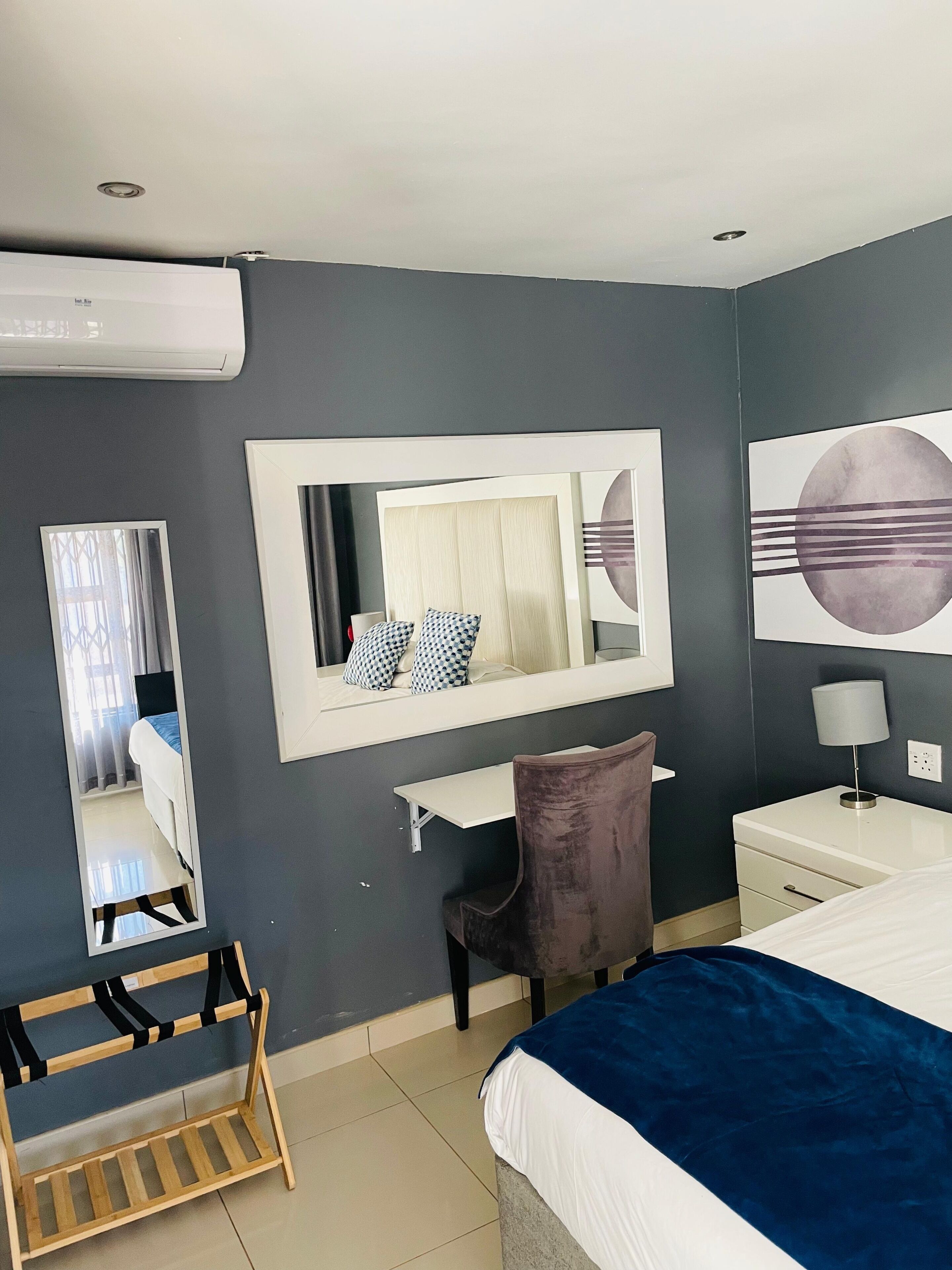 Une chambre d’hôtel moderne équipée d’un lit, d’un bureau, d’une chaise, d’un miroir, d’une table de chevet et d’un climatiseur fixé au mur.