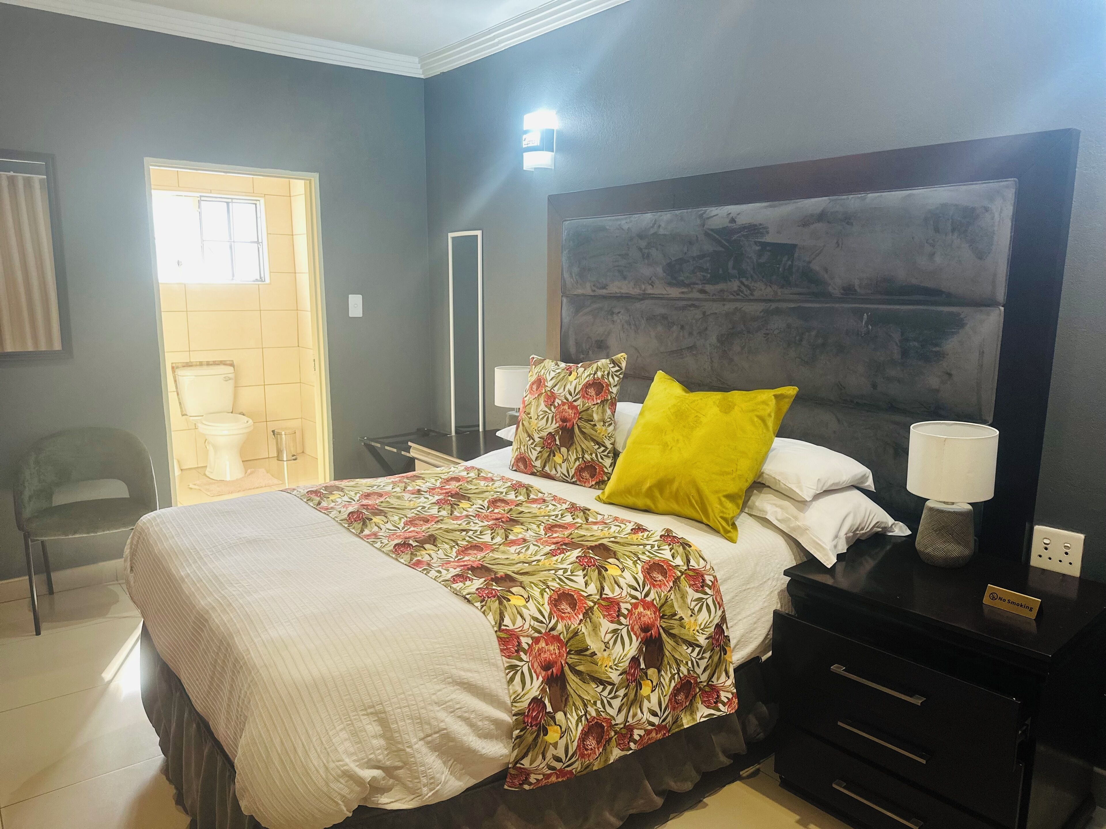 A hotel room with a bed, a nightstand, a chair, a bathroom with a toilet, and a large abstract painting on the wall.