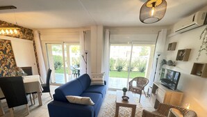A living room with a blue sofa, a dining table with chairs, a TV on a wooden cabinet, and a sliding glass door leading to a patio. - Appartement 2 Piscines, 100m de la Mer, 4/6 Personnes (Santa-Maria-Poggio)