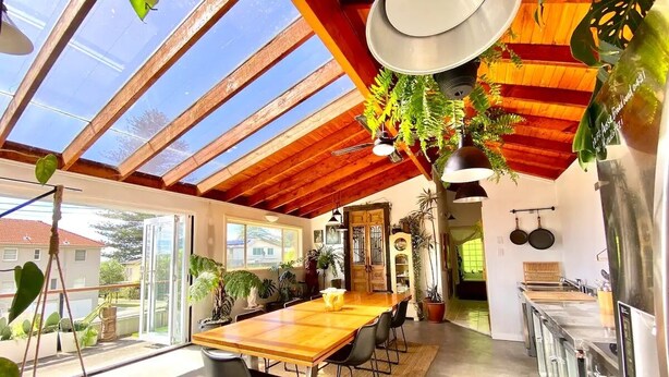 Un vaste espace dédié à la salle à manger, avec une grande table et des chaises en bois, un coin cuisine équipé d’appareils modernes, et une vue sur l’extérieur grâce à un plafond en verre.