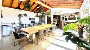 Une cuisine spacieuse avec un plafond en bois, une longue table à manger et plusieurs chaises. On y trouve également des plantes en pot et une statue, situées sur le côté droit.