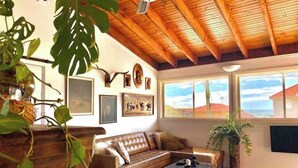 Un salon avec un plafond en bois, un canapé en cuir marron, une table basse et une grande fenêtre donnant sur des immeubles.