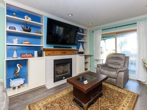 A living room with a fireplace, television, and a grey armchair. - Palm Tiki by Miss Kitty’s (Rockport)