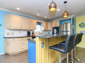 A kitchen with light wooden cabinets, a granite countertop, a stainless steel refrigerator, and a bar area with stools. - Palm Tiki by Miss Kitty’s (Rockport)