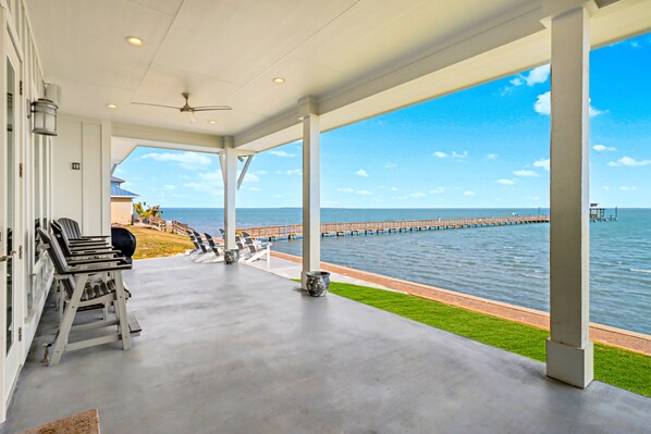 A covered outdoor area with a view of the sea, featuring a concrete floor, white columns, and outdoor seating. - Bayfront water-view home with outdoor pool, lovely patio, hammock, fishing dock (Rockport)