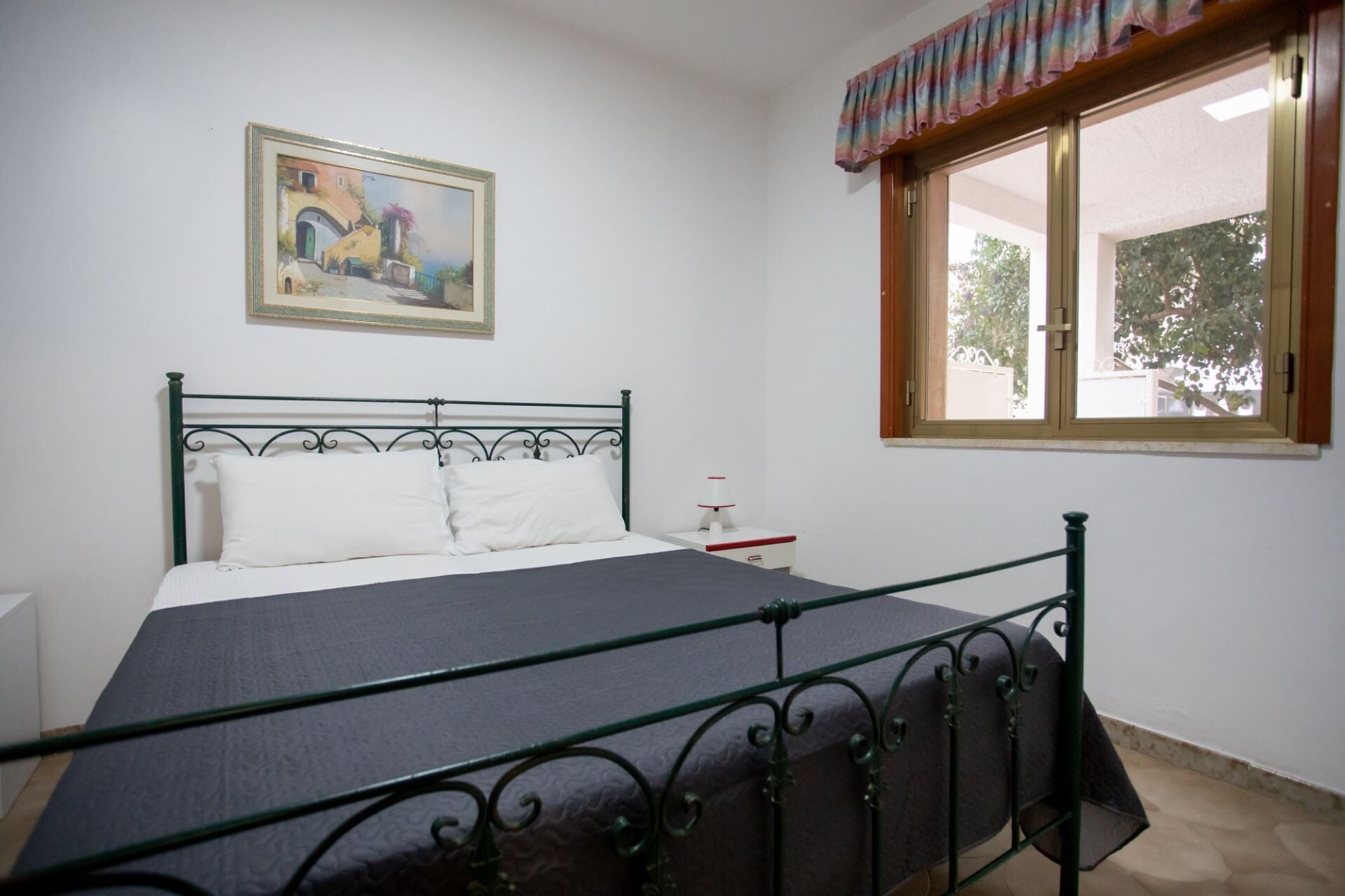 A bedroom with a bed, a nightstand, a lamp, a window with a view, and a painting on the wall.