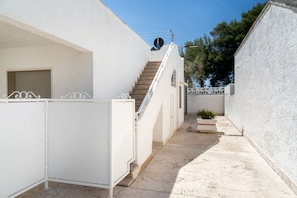 Un patio estrecho con paredes blancas, una escalera que lleva a una puerta, una antena parabólica y una jardinera con plantas.