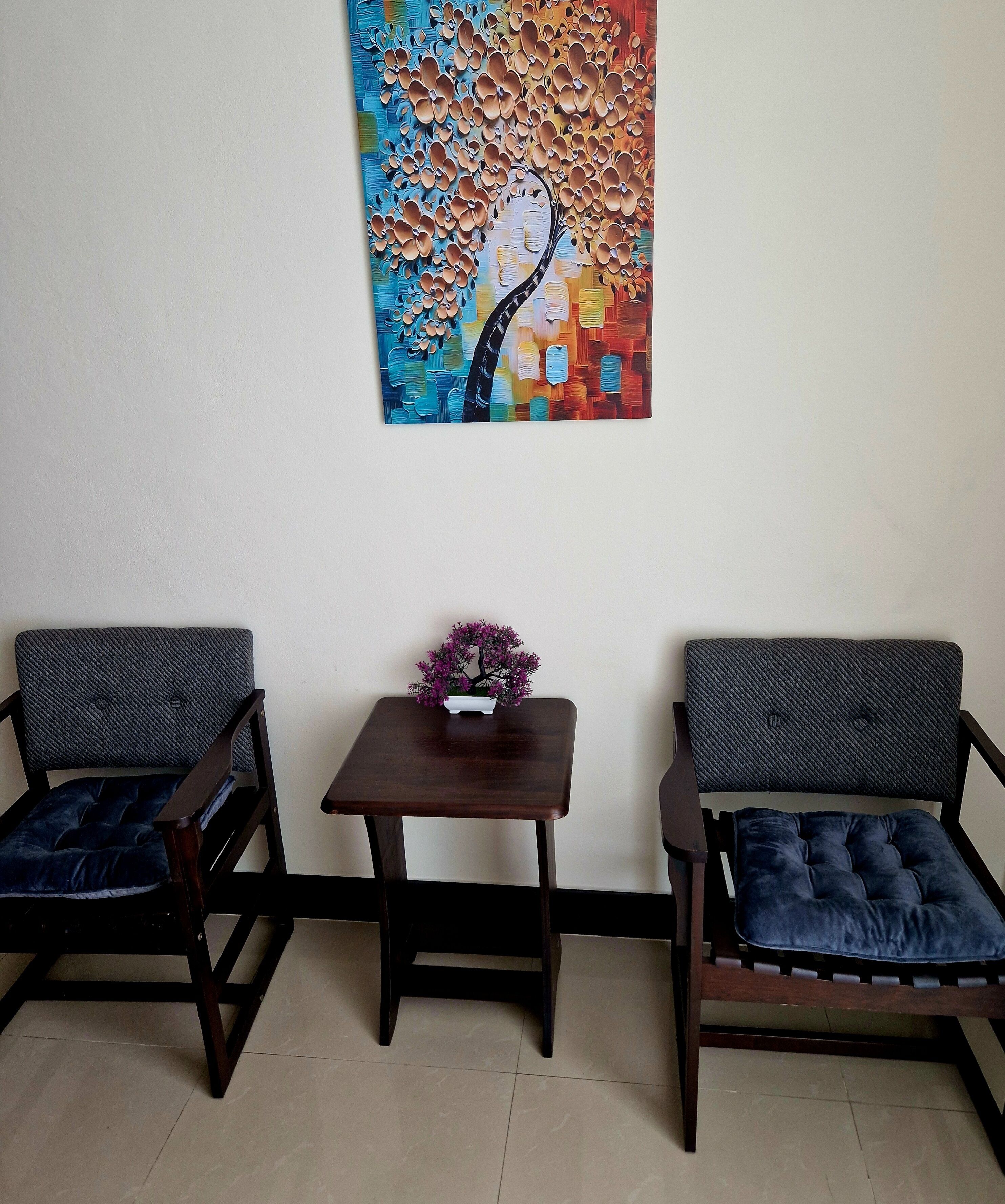 Two wooden chairs with cushioned seats and backs, a small wooden table with a vase of flowers, and a colorful abstract painting on the wall.