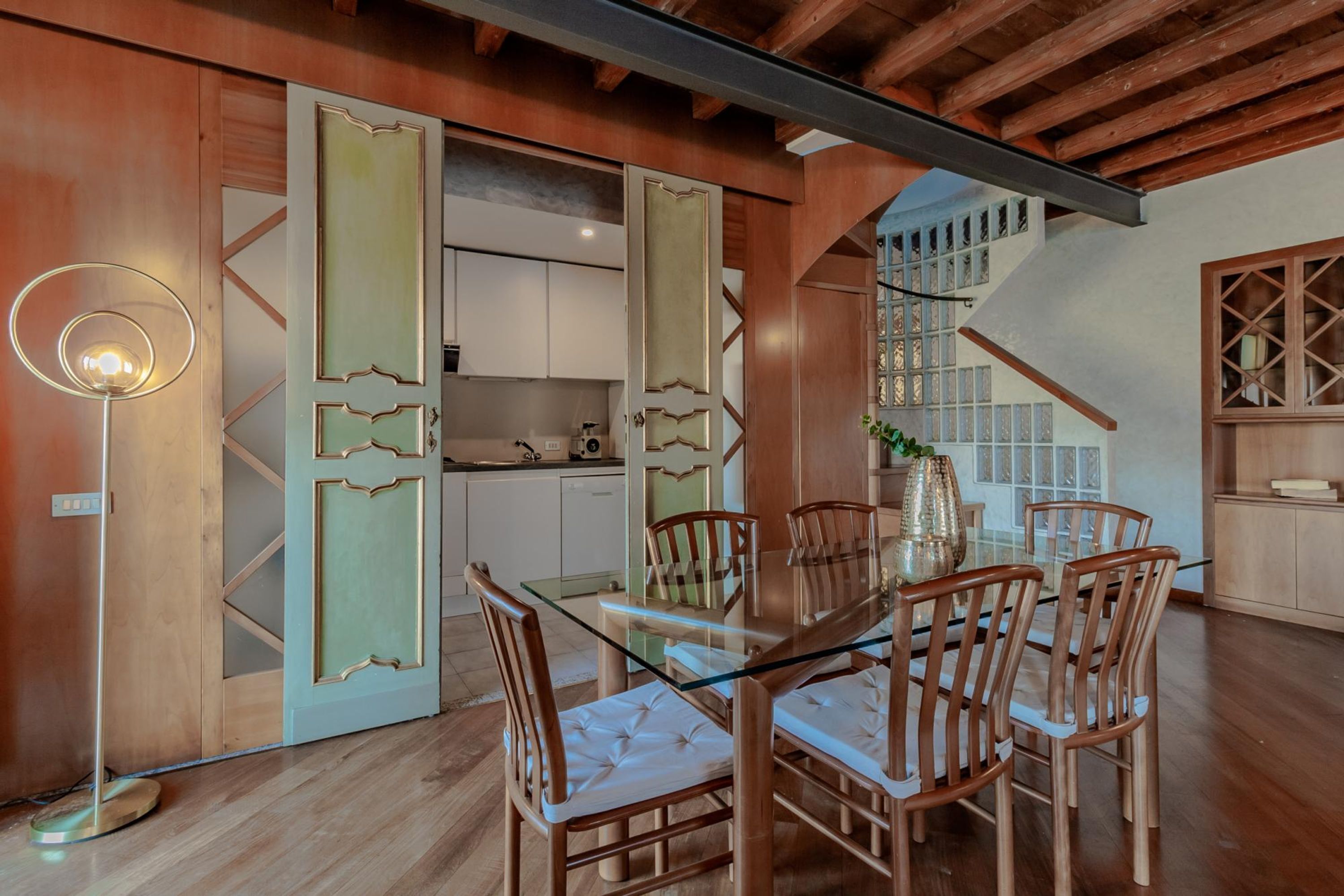Un coin repas avec une table en verre et des chaises en bois, une cuisine moderne en arrière-plan et un escalier agrémenté d’une rampe décorative.