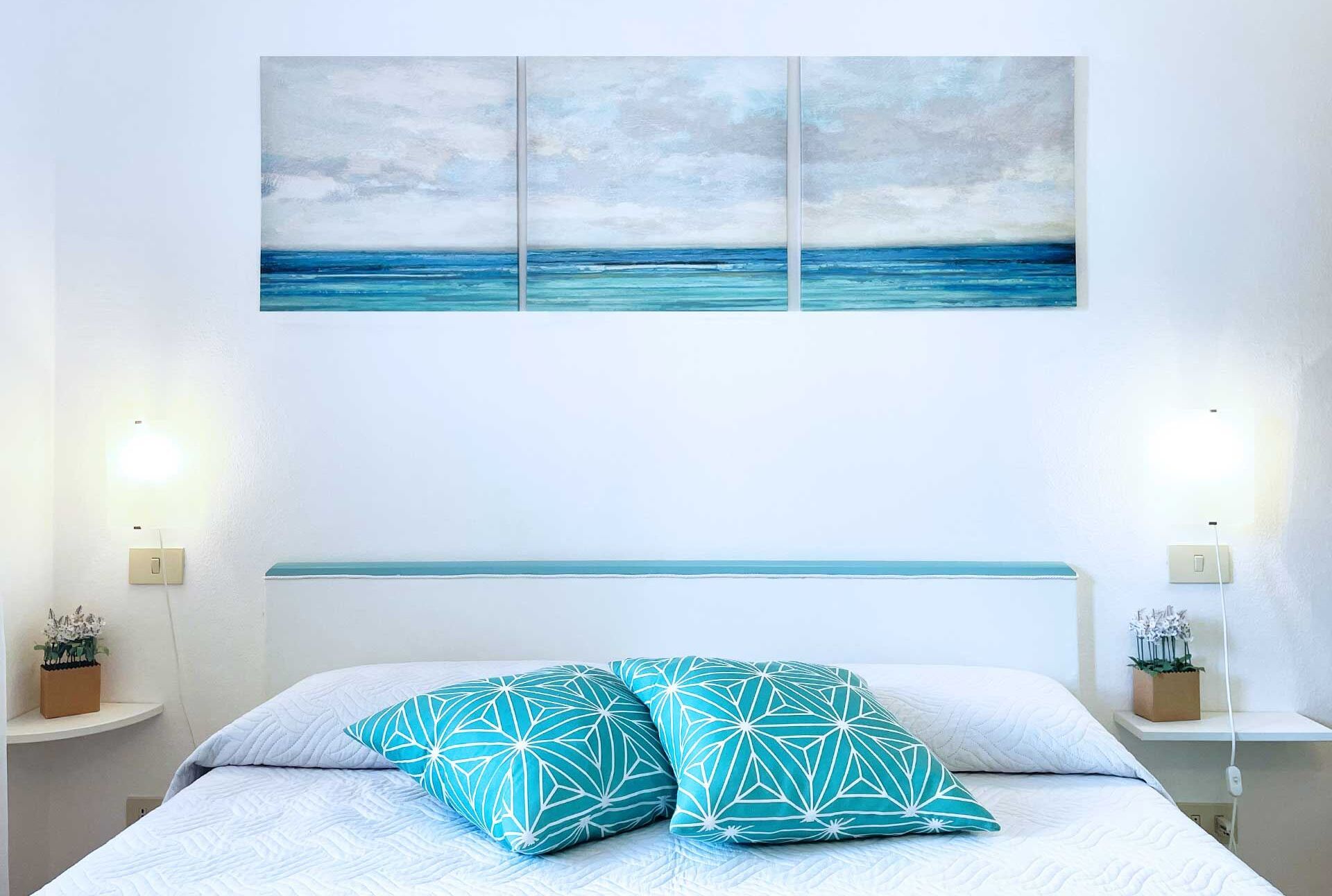 A bedroom with a bed, two nightstands, a wall-mounted shelf, and a painting of a seascape.