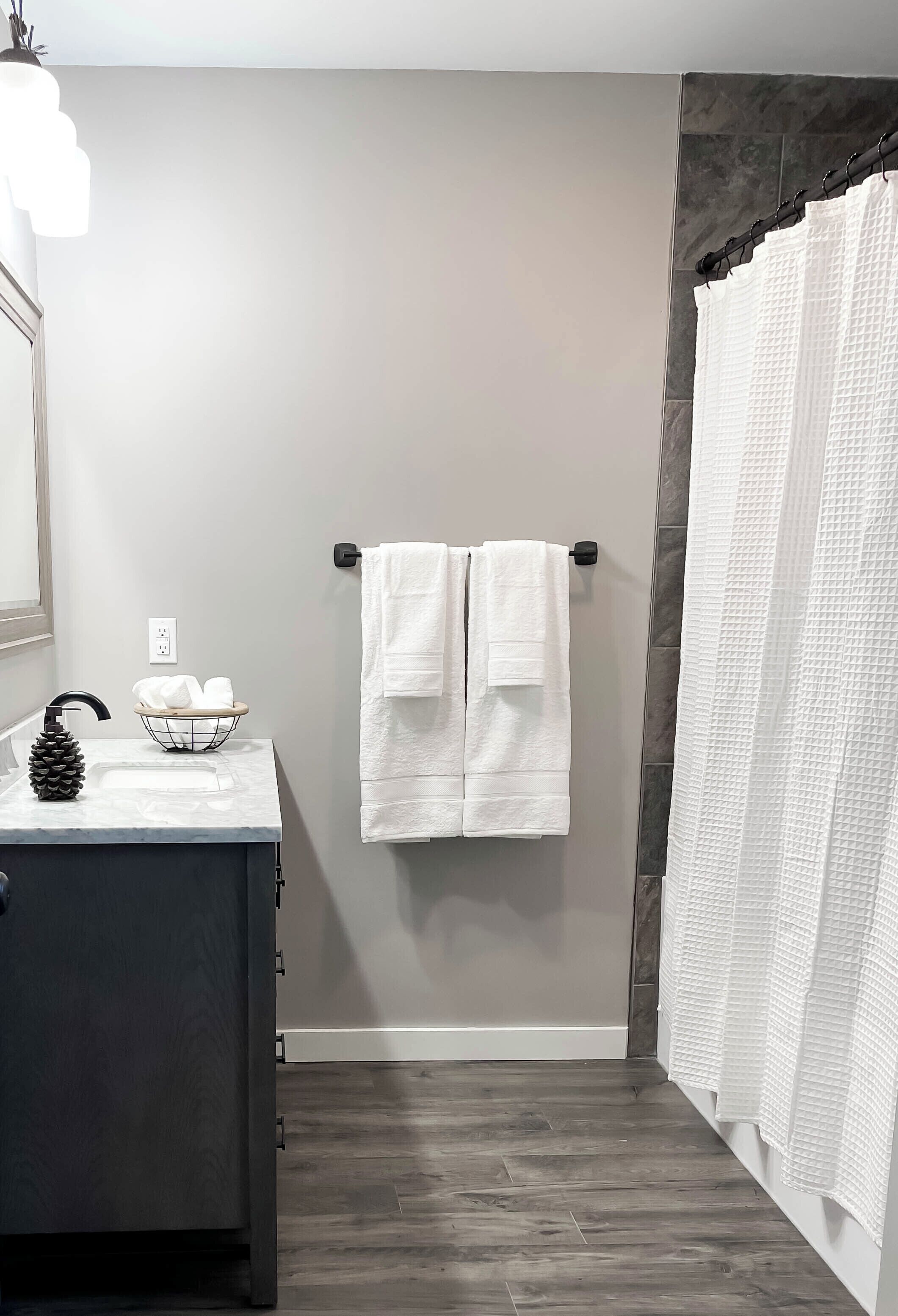 A bathroom with a dark wood floor, a white sink with a pine cone decoration, a towel rack with two white towels, and a white shower curtain.
