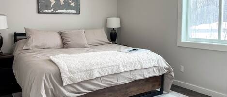 Un dormitorio con cama, dos mesitas de noche, un mapa mundial en la pared, una ventana con vista a árboles y un plaid estampado sobre la cama.