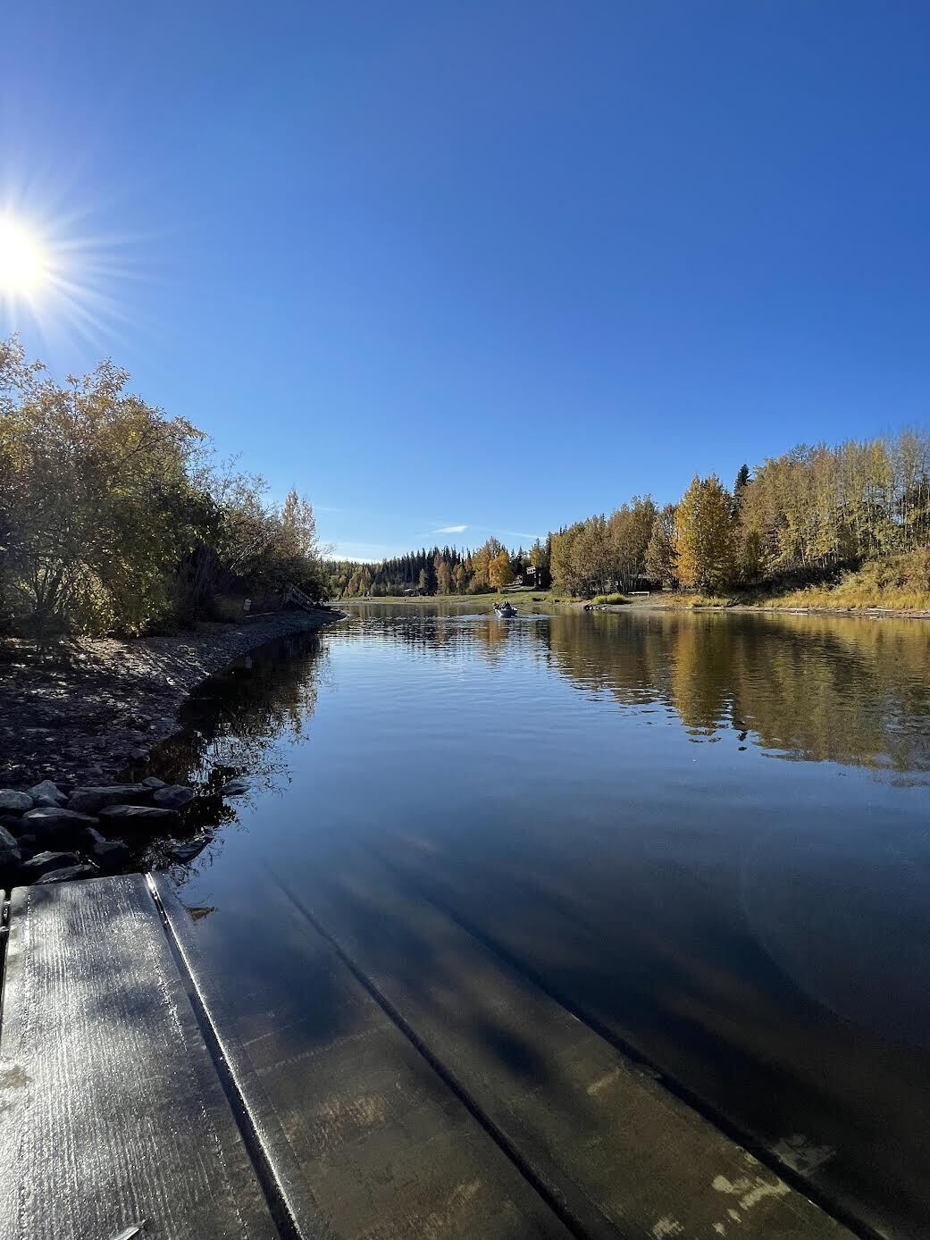 Un río tranquilo con un muelle a la izquierda, rodeado de árboles y un cielo despejado.