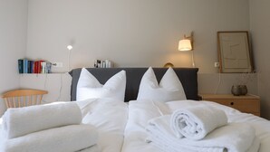En pænt redt seng med hvide sengetøj og puder, et natbord med lampe, en lille træstol og en hylde med bøger og toiletartikler.
