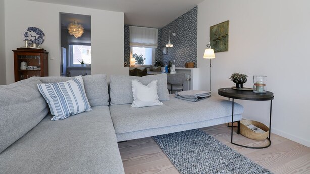 En moderne stue med en grå hjørnesofa, et lille rundt bord med et stearinlys og en grå måtte.
