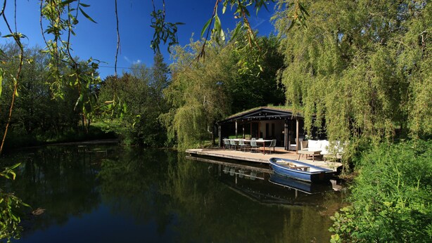 Un chalet paisible situé au bord de la rivière, avec un quai et un petit bateau, entouré d’une végétation luxuriante et d’arbres.